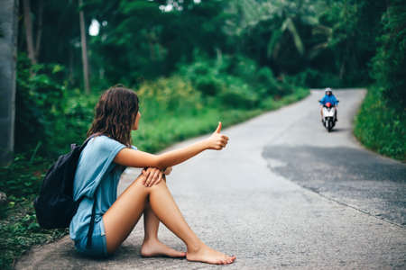 Young beautiful woman hitchhiking sitting on road barefoot. Travel conceptの写真素材
