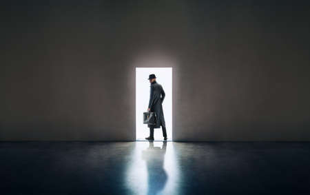 Man silhouette in hat and raincoat standing in the light of opening door in dark roomの写真素材