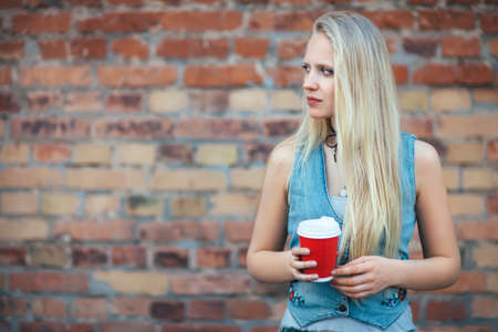 Beautiful blonde woman holding cup of coffee on brick wall background. Urban lifestyleの写真素材