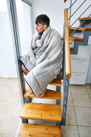 Young handsome man covered with blanket drinking hot coffee sitting on stairs at homeの写真素材