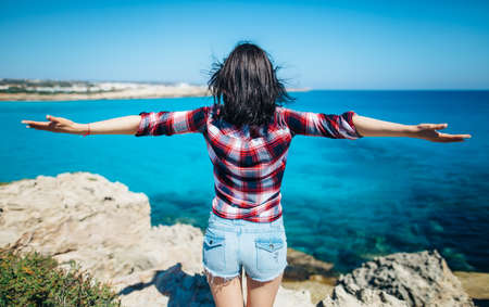 Back view of young woman with wind in her hair raising her hands on top of cliff above beautiful sea. Relax concept.の写真素材