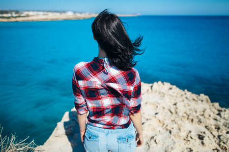 Back view of young woman with wind in her hair standing on cliff and enjoying beautiful sea view. Relax conceptの写真素材