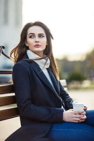 Thoughtful beautiful woman with cup of coffee sitting on a bench in city street. Relax conceptの写真素材