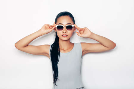 Young beautiful Asian woman standing in summer grey clothes and sunglasses. Attractive tanned girl in casual style. Half body conceptual portrait on theme of fashionable people, accessories, eyewearの写真素材