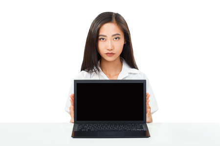 Serious Asian young woman sitting at table with laptop. Black netbook screen with copy space Portrait of female model isolated on white backgroundの写真素材