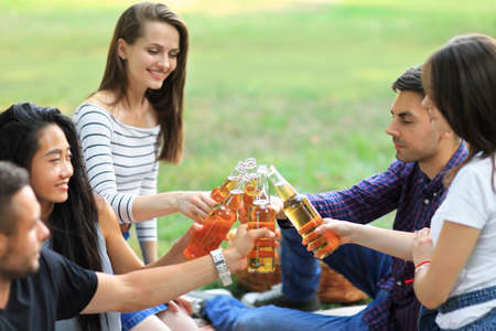 Cheerful carefree group of friends clinking bottles drink on background of blur sunny forest. Young men and women students having fun in warm company outdoors. People enjoying partyの写真素材