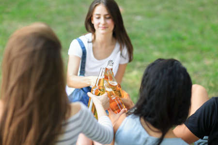 Cheerful carefree group of friends clinking bottles drink on background of blur sunny forest. Young...