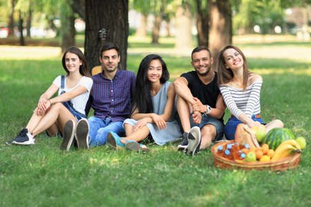 Group of friends sitting in park on grass and looking at camera. Men and women rest on nature with guitar and enjoy life. Young beautiful people outdoors on sunny dayの写真素材
