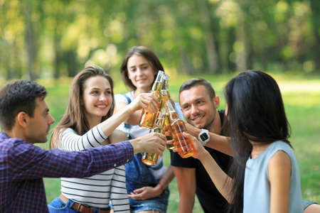Cheerful carefree group of friends clinking bottles drink on background of blur sunny forest. Young men and women students having fun in warm company outdoors. People enjoying partyの写真素材