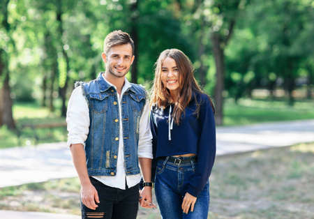 Pair of young people standing in park, hold hands and looking at camera. Romantic meeting of lovers, man and woman in casual style dressed. Gentle date between two lovers in bosom of natureの写真素材