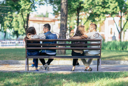 Two lovers couples sitting on different ends of one bench. Young happy men and women communicate...