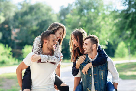 Two loving couples piggyback having fun outdoors. Girls sitting on back of guys smiling. Young women and men playing in park, and looking at each otherの写真素材