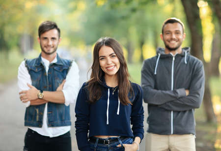 Beautiful young woman and two men with arms crossed behind her. Half body portrait of three people on nature. Girl and guys looking at camera. Focus on female model. Lady makes the choice conceptの写真素材
