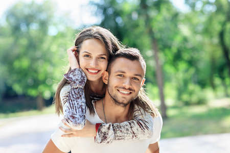 Outdoors portrait of lovers happy young man and woman looking at camera. Smiling girl piggyback of her boyfriend. Love, youth, relationship concept photo on nature background on sunny dayの写真素材
