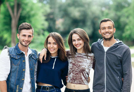 Four young cheerful friends are walking on warm day. Two Caucasian men and two women having fun together outdoors. Half body portrait of smiling people. Models looking at cameraの写真素材