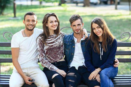 Four women and men sitting on bench in embrace. Happy stylish young friends posing on bench outdoors, smiling and looking at camera. Beautiful healthy people conceptの写真素材