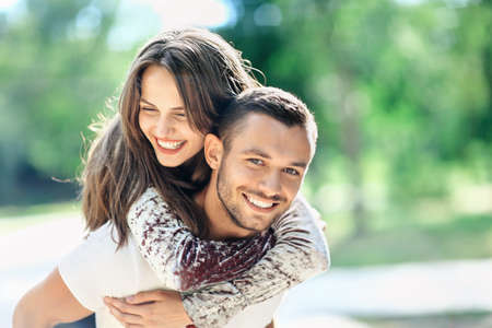 Outdoors portrait of lovers happy young man and woman looking at camera. Smiling girl piggyback of her boyfriend. Love, youth, relationship concept photo on nature background on sunny dayの写真素材