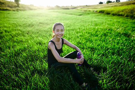 Oriental sports girl on grass with bottle looking into camera and smiling. Woman after run sitting. Concept of outdoor sports, healthyの写真素材