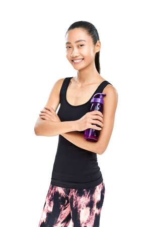 Asian woman in sportswear with bottle of water. Fitness girl resting and standing with arms crossed and sporting plastic bottle. Isolated portrait of female model bright smiling and looking at cameraの写真素材