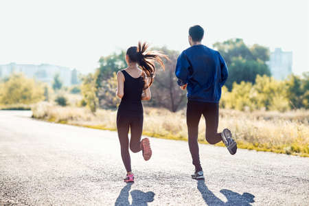 Active sporty woman and man jogging on sunny day in park. Couple in tracksuits running along the road. Healthy lifestyle, sport, healthcare concept. Full-length portrait of two people, back viewの写真素材