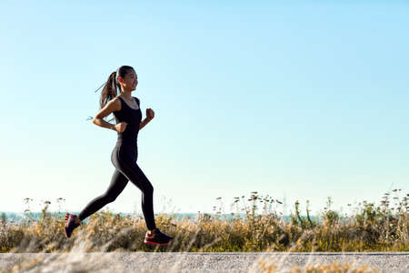 Young athletic woman jogging on rural road in early morning. Sporty girl in black training clothes running toward the sun. Photo with copy space on blue sky background. Full-length portraitの写真素材