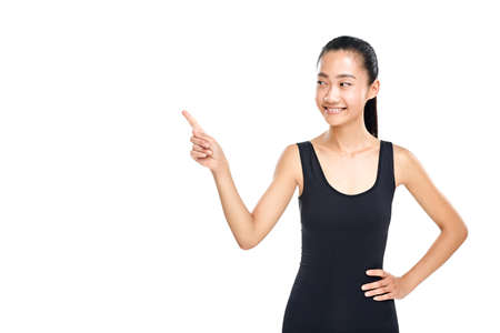 Asian woman foxy smiling and pointing finger at something. Slender girl in sporty black tanktop. Presentation, explanation, instruction concept. Half body portrait isolated on white backgroundの写真素材