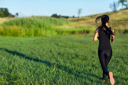 Back view of woman in black sportswear running. Japanese runs with flying hair and casts shadow over green grass. Active sport conceptの写真素材