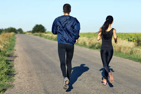Active sporty woman and man jogging on sunny day in park. Couple in tracksuits running along the road. Healthy lifestyle, sport, healthcare concept. Full-length portrait of two people, back viewの写真素材