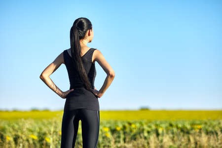 Sports woman standing in field and admiring nature on clear fine day. Half body portrait of girl in black sportswear with hands on hips, back view. Athlete resting after joggingの写真素材