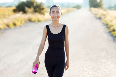 Asian athletic woman walking on rural road and carrying water bottle in her hand. Smiling slender girl with oriental appearance in black tracksuit going on countryside workout or joggingの写真素材