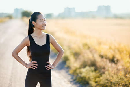 Slim athletic Asian woman resting on rural road in sunny day. Half body portrait of girl in black sportswear with hands on hips, model looking thoughtfully into the distance and enjoying the sunの写真素材