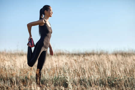 Young Asian woman stretching legs on field on warm day. Full-length portrait in profile of sporty...