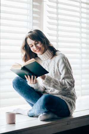 Happy beautiful woman reading book sitting on window sill at home. Relax conceptの写真素材