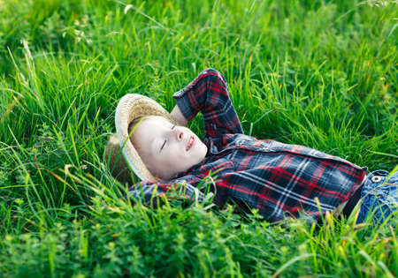 Handsome little boy lying on grass on back, hands behind head. Little dreamer lies in meadow with hat, eyes closed. Child sleeping and smiling in nature outside city. Carefree pastime, vacationの写真素材