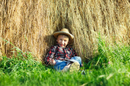 Happy boy in straw hat sitting in haystack, outdoors, looks camera. Smiling child relax haystack on green grass under the hot sun. Young man spends holidays in village outside cityの写真素材