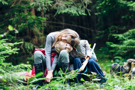Hugging children are sitting in forest glade, their heads inclined. Brother and sister are playing in clearing surrounded by trees and with dog. Family bonds の写真素材