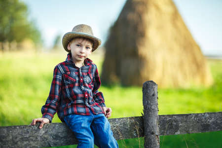 Child sits on wooden fence against picturesque haystack, close up. Young boy in casual clothes resting on fence in village. Country vacations conceptの写真素材