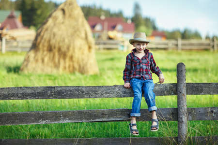 Little boy in hat sitting on fence against of haystack. Child on a hedge against backdrop of rural landscape. Young cowboy smiling shyly and looking at camera. Village, country trip, rest, conceptの写真素材