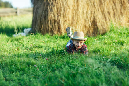 Handsome little boy lies on grass on background of haystack. Child rests in nature, lies on his stomach and propps head with hands. Portrait of young man outdoors. Holidays, vacations, weekend conceptsの写真素材