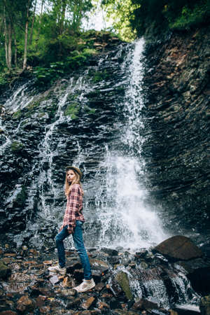 Portrait of young girl near waterfall. Young lady poses against background of water falling from the cliff, overgrown with grass, bushes and forest. Photoshoot in wild outside cityの写真素材