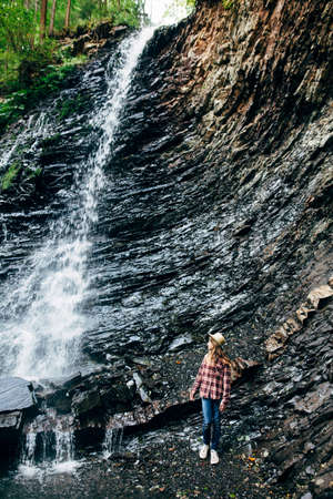Portrait of young girl near waterfall. Young lady poses against background of water falling from the cliff, overgrown with grass, bushes and forest. Photoshoot in wild outside cityの写真素材