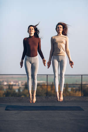 Two beautiful young women synchronously jump on the roof outdoors. Girlfriends are engaged in acrobatics and yoga. Frozen jumping womenの写真素材