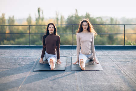 Two beautiful women sitting on a hanumanasana or monkey pose on mats on the roof, outdoors....