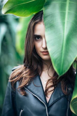 Fashion portrait of beautiful mysterious woman hide behind big leaf, close up. Girl with half closed face in greenhouse