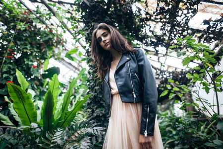 Fashion portrait of young stylish woman in pink dress and leather jacket posing on garden greenhouse background. Beauty conceptの写真素材