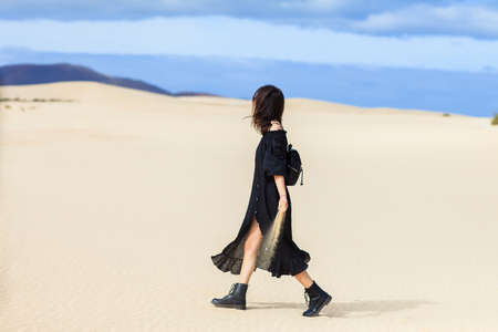 Young lonely woman in long black dress walking in desert holding sand let it running through hands. Time running, lost, mystery conceptの写真素材