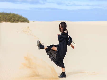 Beautiful woman kicking sand in desert on Spain. Girl in long black dress and with backpack having fun and enjoying summer vacationの写真素材