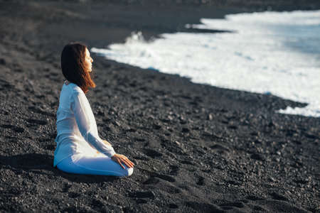 Young woman meditating in Lotus Pose in unique black sand beach Playa Bermeja, Lanzarote, Canary Islands, Spain. Padmasana. Relax conceptの写真素材