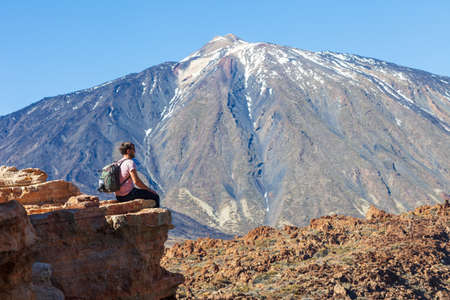 Traveler man with backpack sitting on the edge of rock enjoy beautiful view of volcano El Teide in National park of Tenerife, Canary Islands, Spain. Travel concept.の写真素材