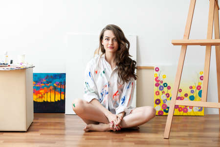 Pretty woman painter sitting on the floor in front of canvas and drawing in her art studioの写真素材